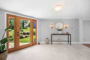 Una habitación con una puerta de madera que da a un jardín, un espejo redondo, una consola decorada con objetos ornamentales y una lámpara en el techo.
