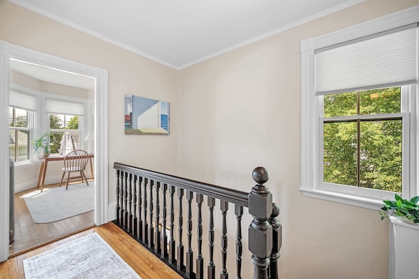 A hallway with a wooden floor, a black railing, and a window with a view of greenery.