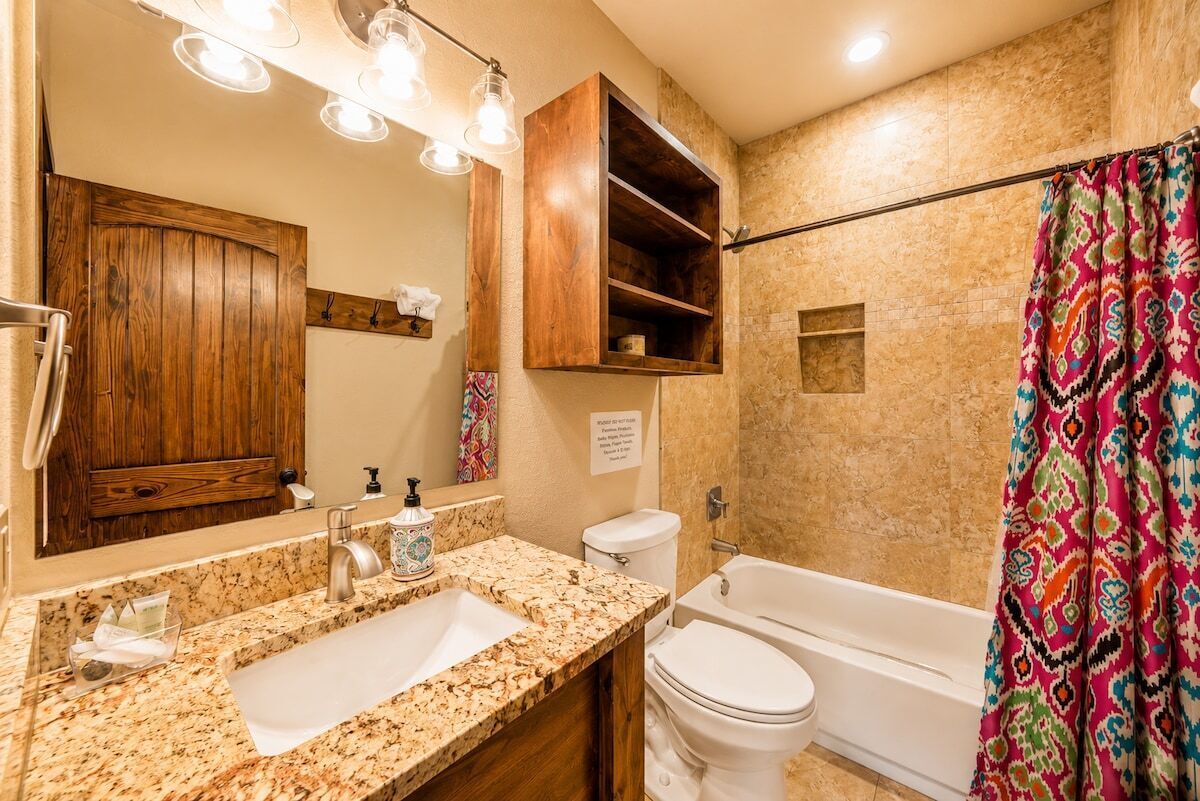 Une salle de bain avec un lavabo surmonté d’un plan de travail en granit, un meuble en bois, un miroir, des toilettes et une baignoire.