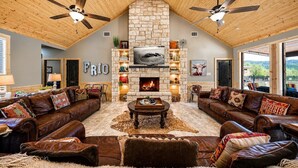 A spacious living room with leather sofas, a stone fireplace, and a large TV screen. - Spacious Lodge w/ Pool & Hot Tub Near Frio River (Concan)