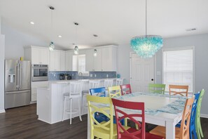 A modern kitchen with white cabinets, a stainless steel refrigerator, and a dining area with colorful chairs. - Canalfront dock and screened porch (Surf City)