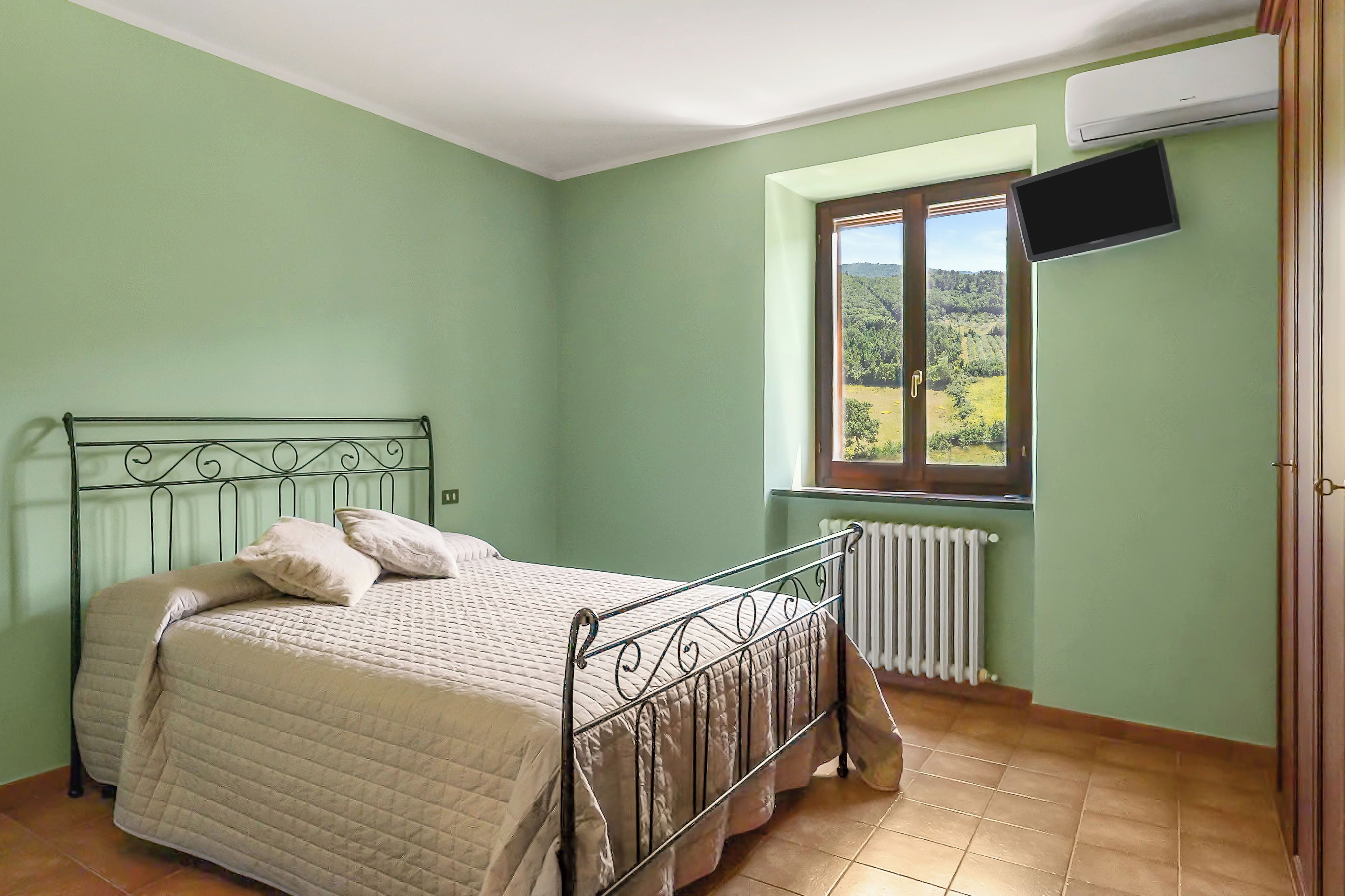 Una camera da letto con un letto, una finestra vista, un termosifone e una TV fissata alla parete.