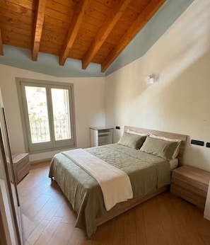 Een slaapkamer met een houten plafond, een bed met een groene dekbedovertrek, een nachtkastje en een raam met uitzicht op groen.
