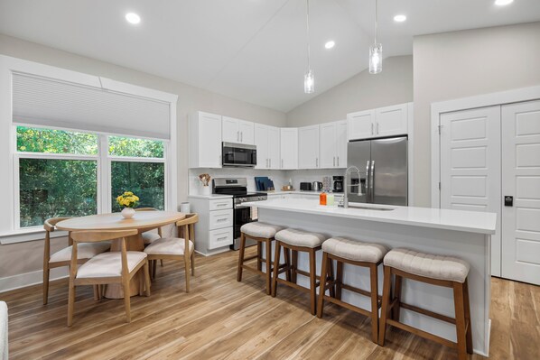 A modern kitchen with white cabinetry, a stainless steel refrigerator, a microwave, and a dining area with a wooden table and chairs.