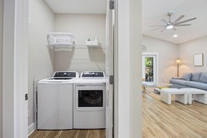 A laundry room with two washing machines and a drying rack, a ceiling fan, and a living area with a sofa and a coffee table. - Cozy 3-bedroom cottage in delightful Pensacola with AC. 10 Mins from the beach. (Pensacola)