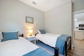 A hotel room with two beds, a nightstand with a lamp, a painting on the wall, and a kitchenette. - Cozy 3-bedroom cottage in delightful Pensacola with AC. 10 Mins from the beach. (Pensacola)