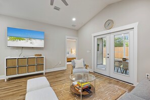 A living room with a flat-screen TV, a sofa, a glass coffee table with board games, and a view through a sliding glass door to an outdoor patio.