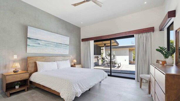 A modern bedroom with a large bed, a ceiling fan, a painting of the ocean, and a view of the outdoor patio. - Luxury Villa + Casita in Mar Vista (Playa Brasilito)
