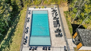 Ein Luftbild eines rechteckigen Schwimmbeckens mit einer umgebenden, mit Fliesen ausgelegten Liegefläche, ausgestattet mit mehreren Liegestühlen und Sonnenschirmen.