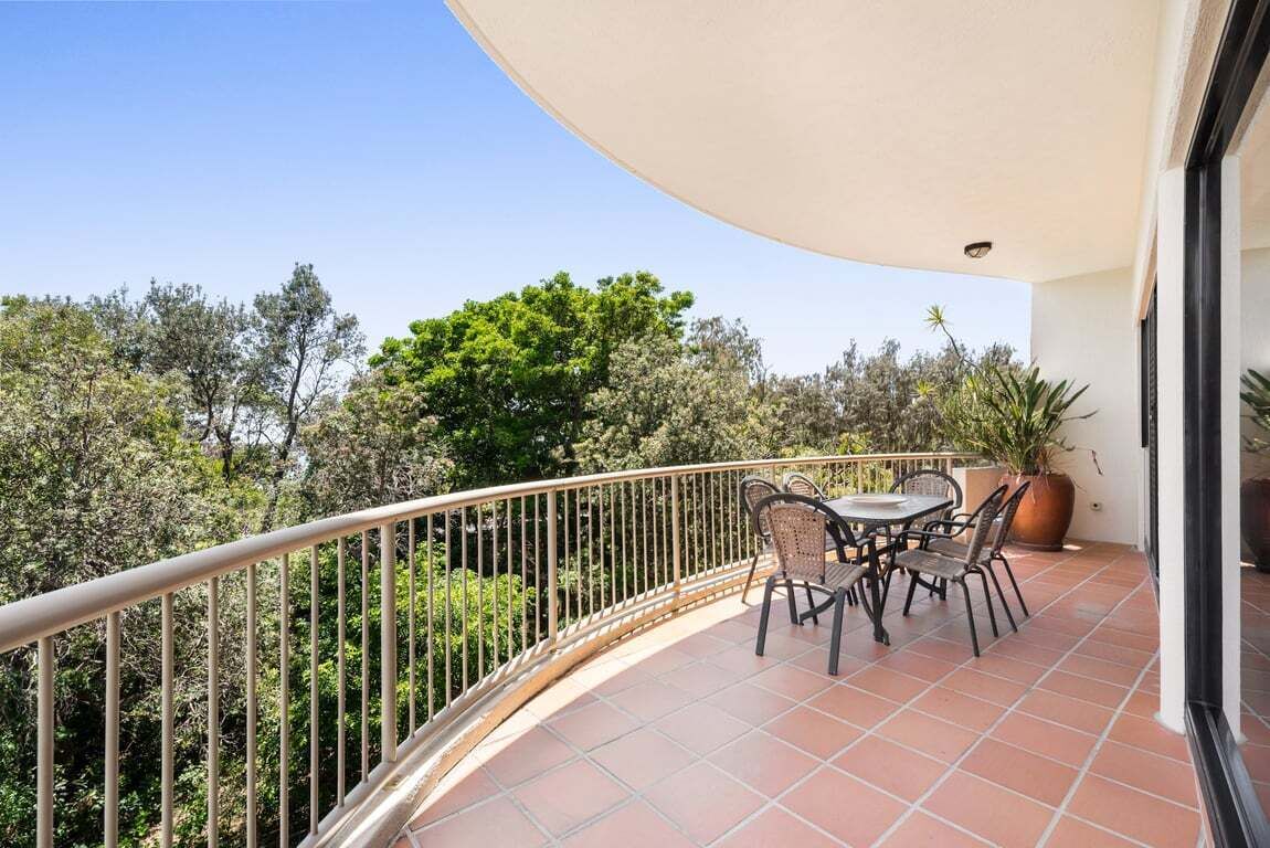 Un balcón con mesa y sillas, rodeado de baranda metálica, con vista a un jardín con árboles y arbustos.