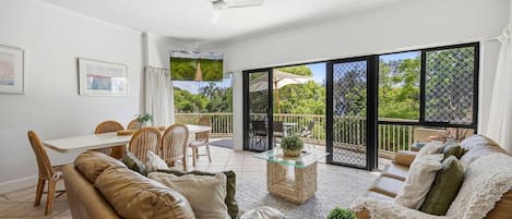 A spacious living area with a sofa, coffee table, and dining set. There is a balcony with a view of greenery.