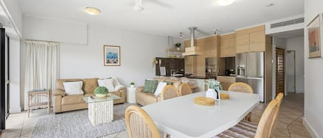 A modern kitchen with white cabinets, a dining area with wicker chairs, and a living room with a sofa and coffee table.
