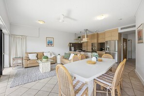 A modern kitchen with white cabinets, a dining area with wicker chairs, and a living room with a sofa and coffee table. - Three-Bedroom Apartment - Exclusive Rooftop Apt 11 (Mooloolaba, Sunshine Coast)