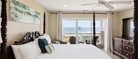 A bedroom with a large bed, a ceiling fan, a view of the ocean, and a balcony with chairs.