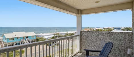 A balcony with a view of the beach and ocean, wooden deck, and a chair.