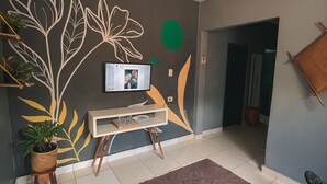 Un salon avec un téléviseur à écran plat posé sur un meuble, une plante en pot, un tapis marron et une personne assise sur un canapé.