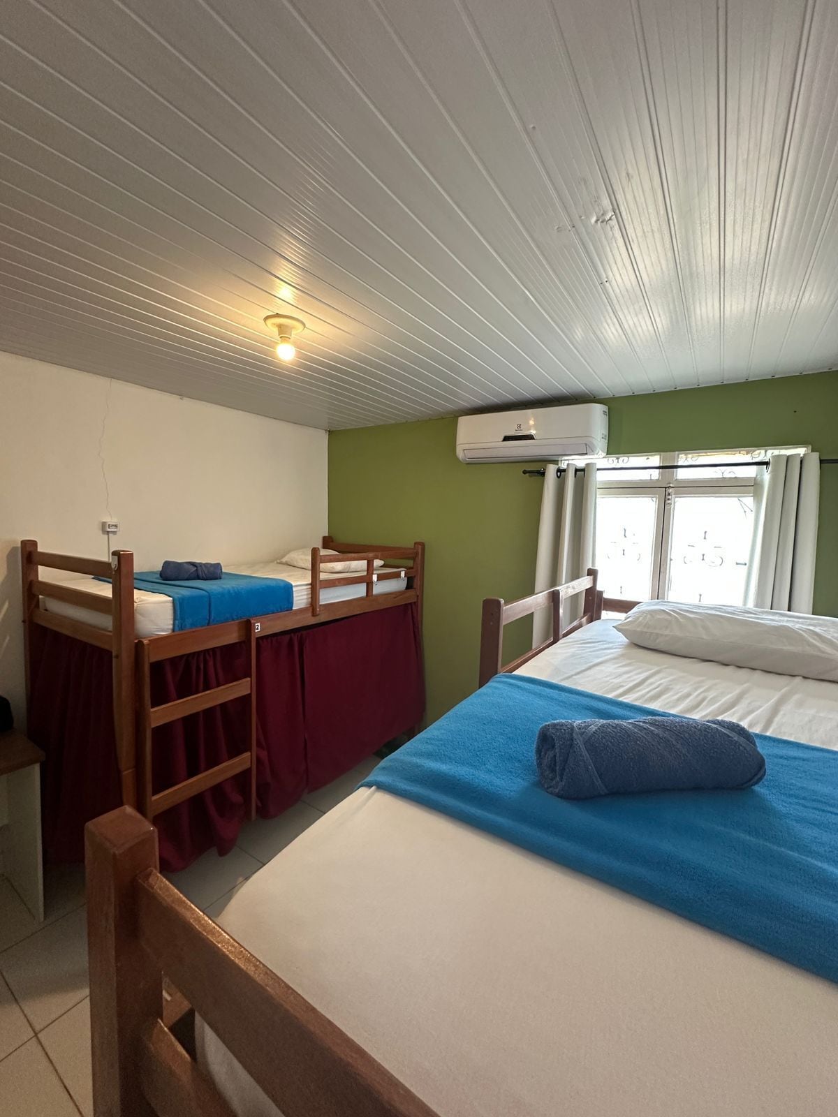 A room with bunk beds, a single bed, an air conditioning unit, and a window with curtains.