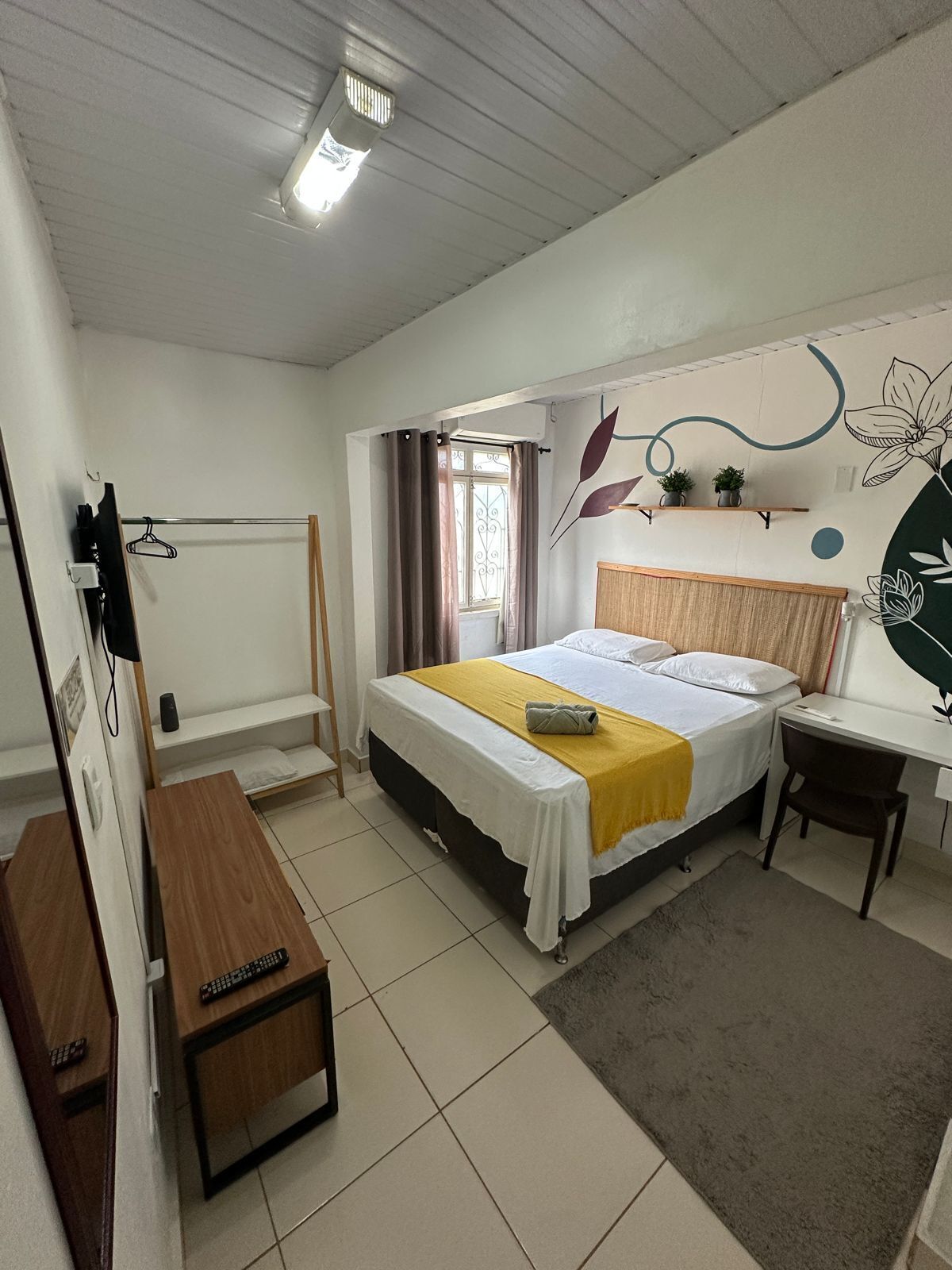 A bedroom with a bed, a desk, a chair, a small table, a radiator, a window with curtains, and wall art.