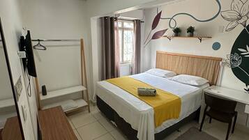 A bedroom with a bed, a desk, a chair, a small table, a radiator, a window with curtains, and wall art.