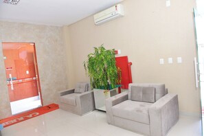A hotel lobby with a reception desk, two armchairs, a potted plant, and an air conditioning unit.