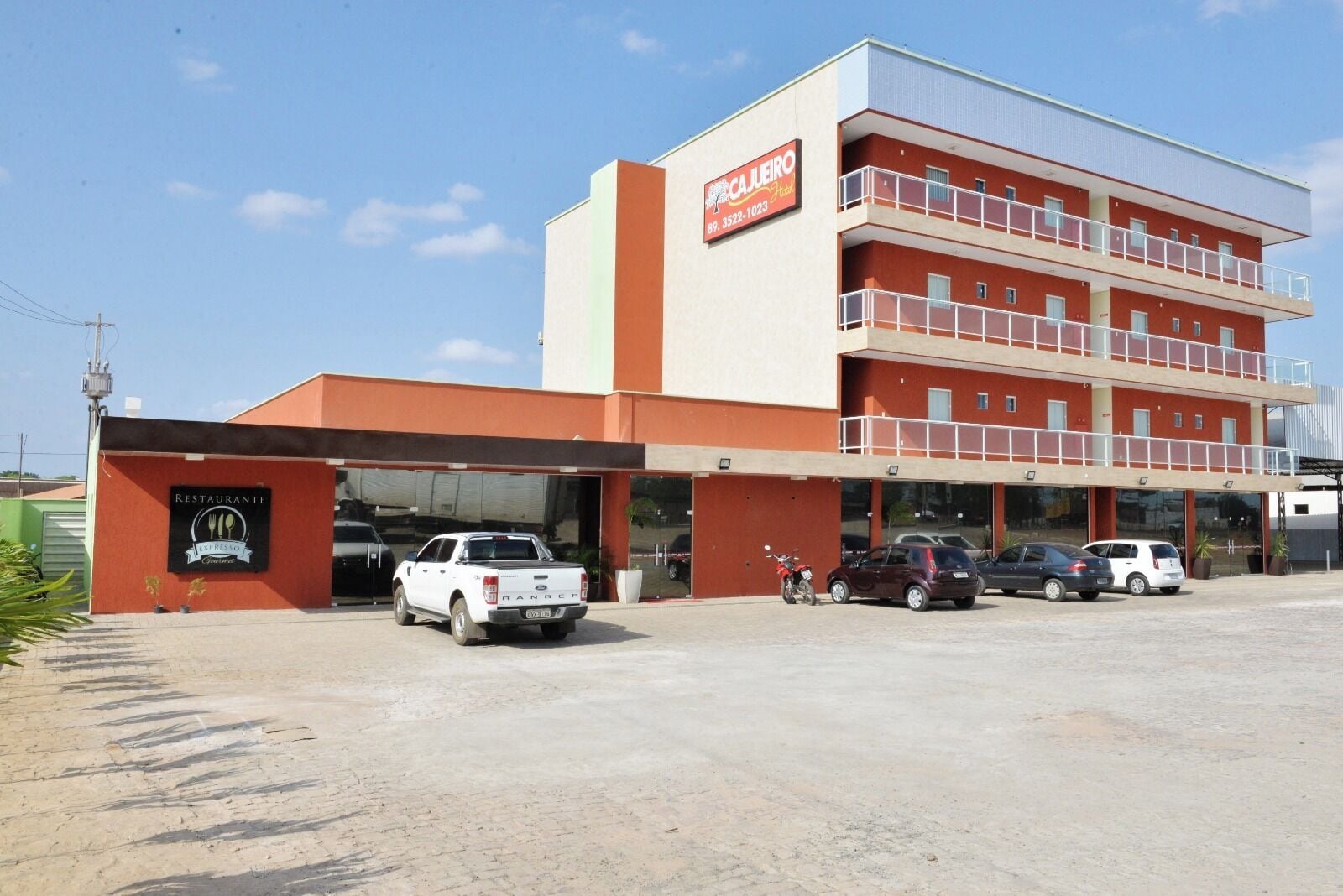 A multi-story hotel with a flat facade, a parking area with several vehicles, and a sign indicating the name and address.