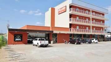 Un hotel de varios pisos con fachada plana, un área de estacionamiento con varios vehículos y un letrero que indica el nombre y la dirección.