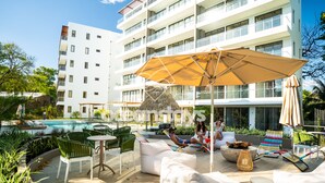 Ein Poolbereich mit Außenmöbeln, einem großen Sonnenschirm und einem Gebäude mit mehreren Balkonen im Hintergrund.