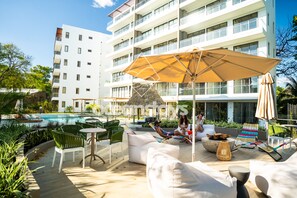 A poolside area with outdoor seating, a large umbrella, and a building with multiple balconies in the background. - The Drifter • 2Bdr • Ocean View & Steps to Surf (El Zonte)