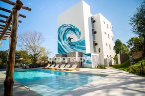 A modern building with a large mural of a surfer riding a wave, a swimming pool with lounge chairs, and a clear blue sky. - The Drifter • 2Bdr • Ocean View & Steps to Surf (El Zonte)