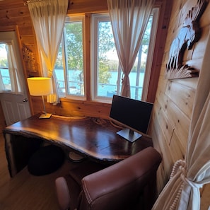 A wooden cabin interior with a desk, chair, and a view of a lake through large windows. - Lil' Bears' Lair (Arnstein)