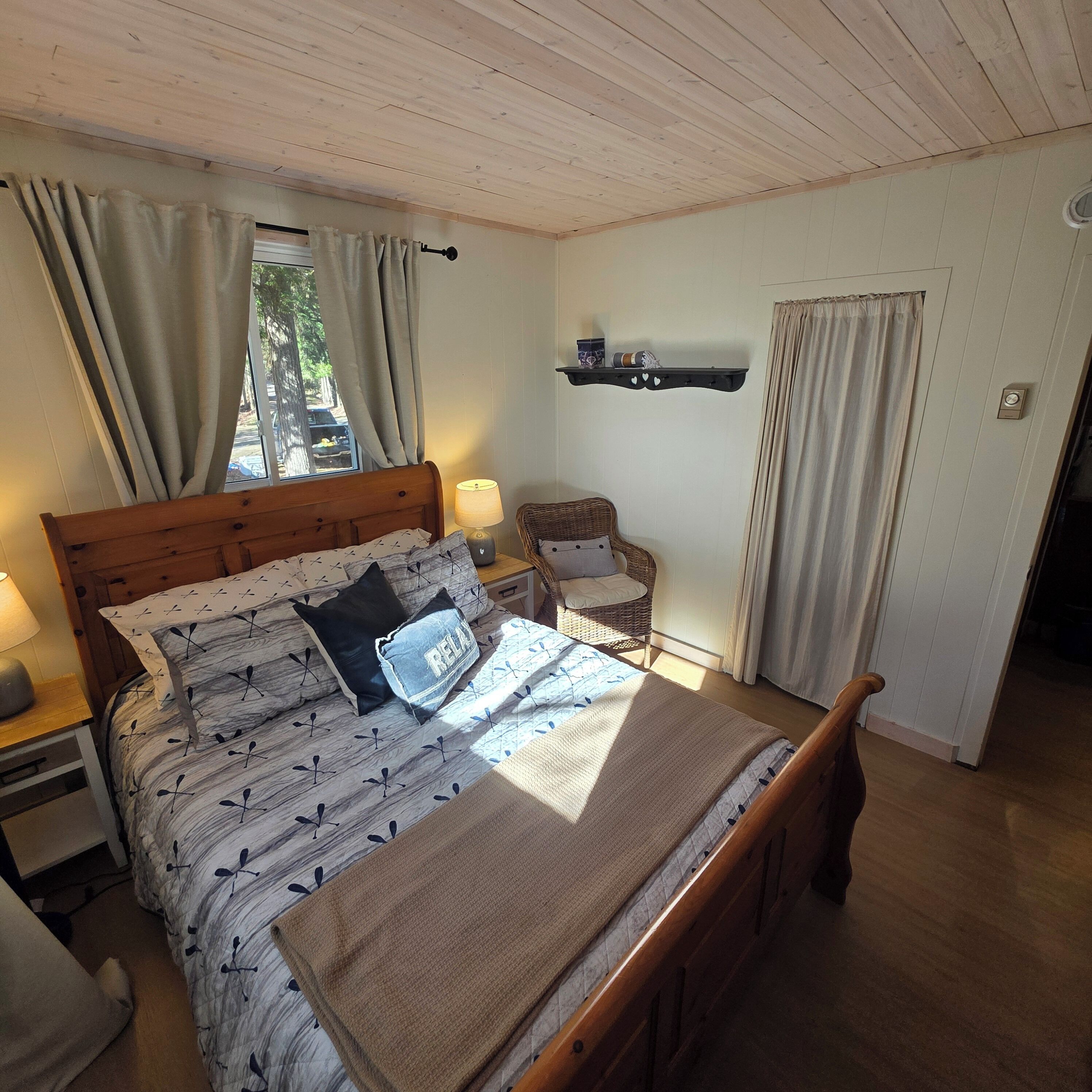 Une chambre à coucher avec un lit en bois, une table de chevet avec une lampe, un fauteuil en osier et une étagère ornée d’objets décoratifs.