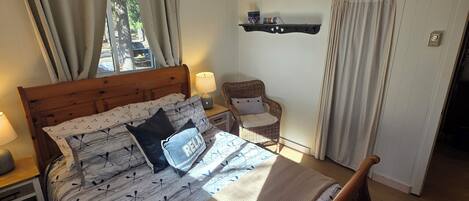Un dormitorio con una cama de madera, una mesita de noche con lámpara, un sillón de mimbre y una estantería con adornos.
