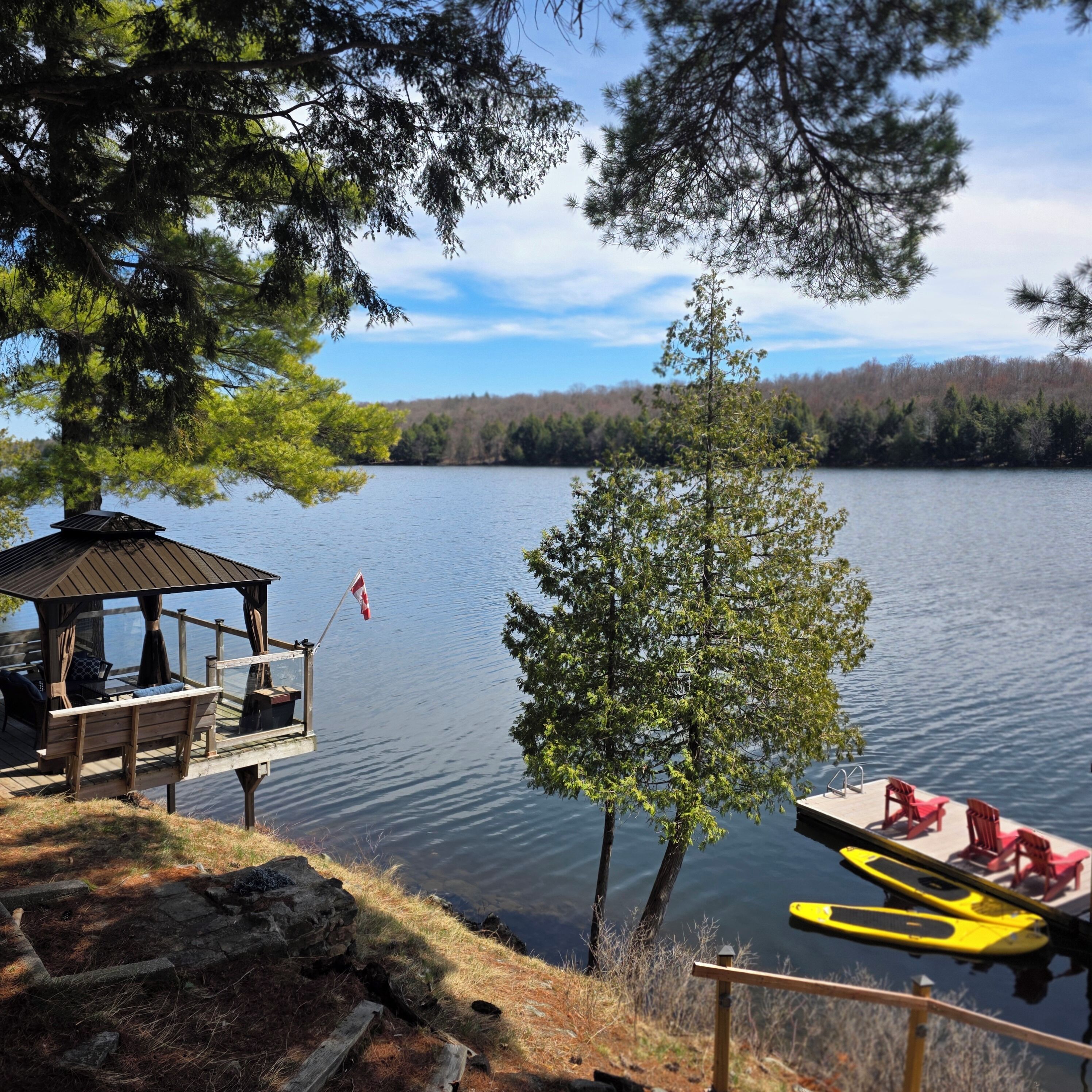 A lakeside wooden deck with a gazebo, a small dock with kayaks, and a tree in the foreground.