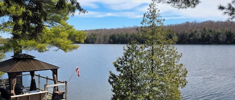 A lakeside wooden deck with a gazebo, a small dock with kayaks, and a tree in the foreground.