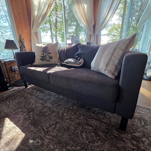 A dark grey sofa with patterned cushions, a wooden floor, and a large window with curtains. - Lil' Bears' Lair (Arnstein)
