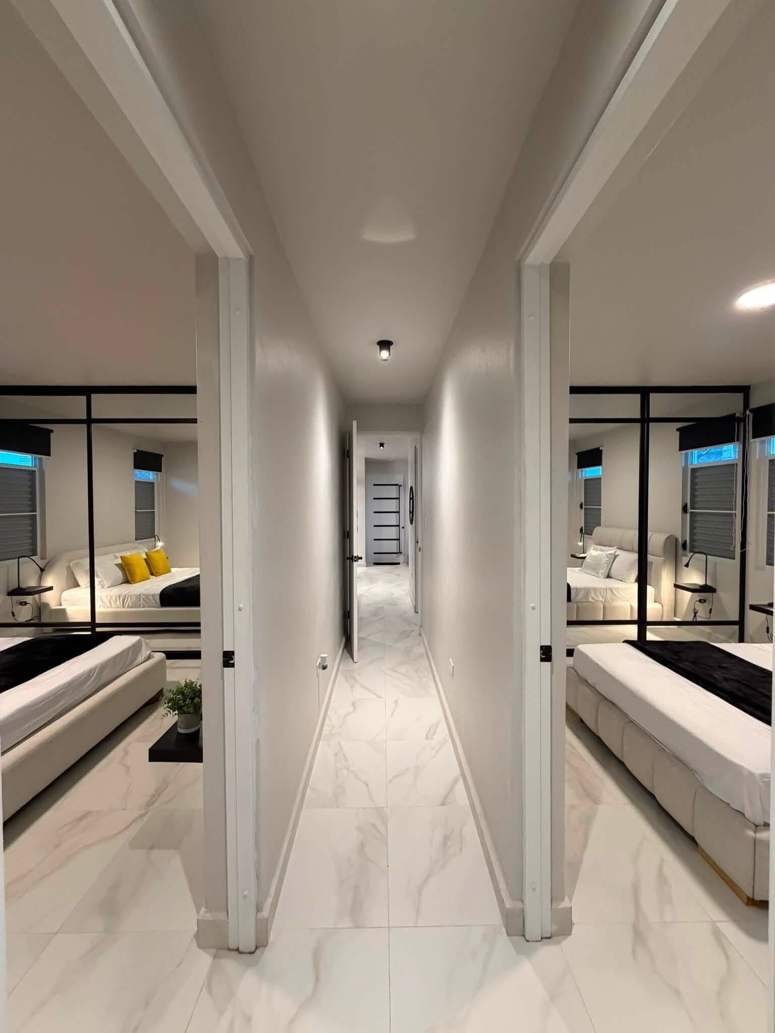 Un pasillo moderno de hotel con dos habitaciones a cada lado, cada una con cama, mesita de noche y lámpara.