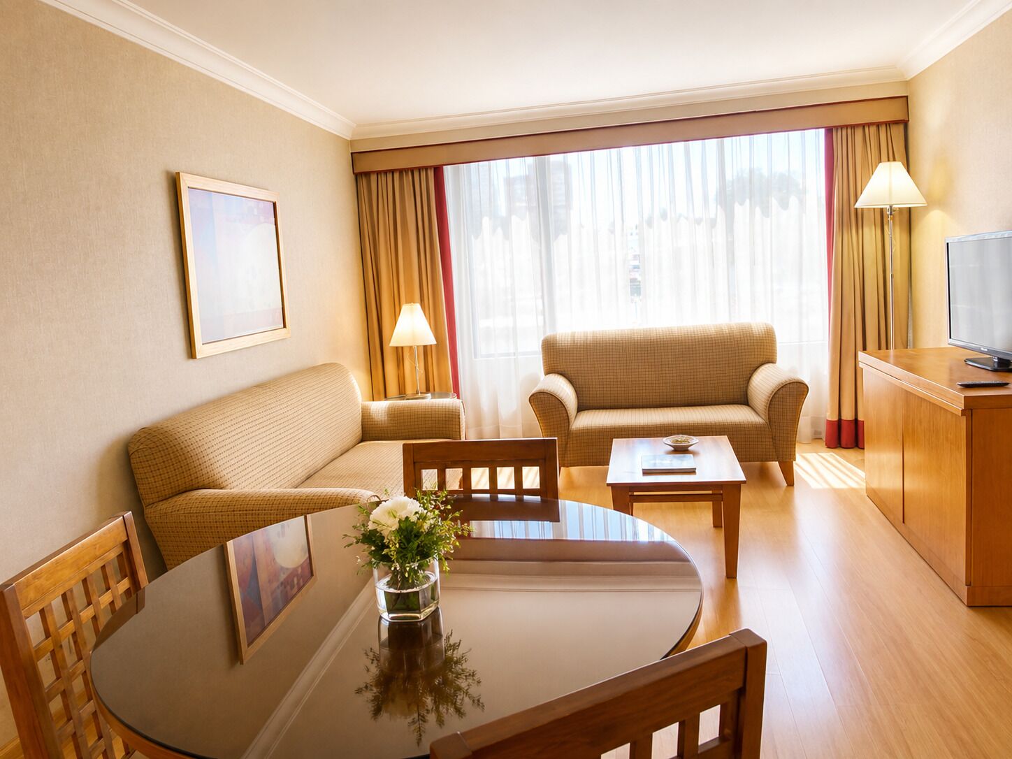 Una habitación de hotel con sofá, una mesa de comedor con sillas, un mueble para televisión y una ventana con cortinas.