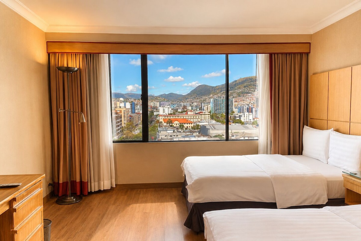 Une chambre d’hôtel avec deux lits, un bureau et une grande fenêtre donnant sur la ville.