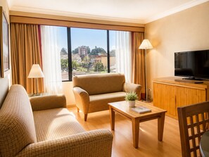 Habitación de hotel con sofá, sillón, mesa de centro, televisor y vistas a la ciudad.