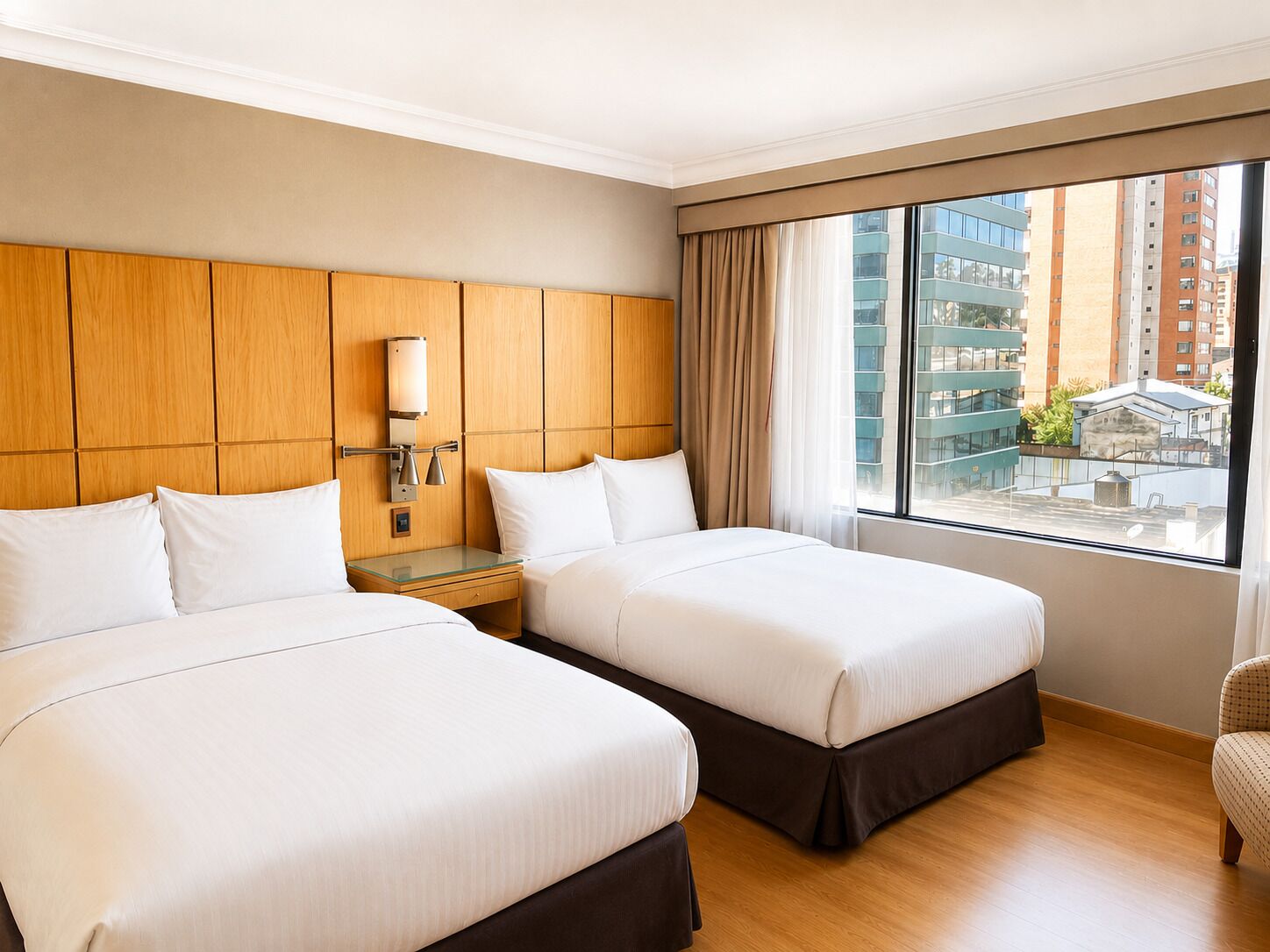 A hotel room with two beds, a wooden headboard, a nightstand with a lamp, and a large window with a view of buildings.