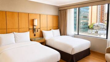 A hotel room with two beds, a wooden headboard, a nightstand with a lamp, and a large window with a view of buildings.