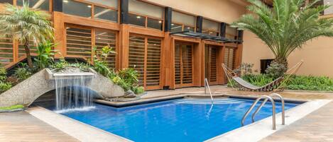 A swimming pool with a waterfall feature, surrounded by a tiled patio, wooden doors, and potted plants.