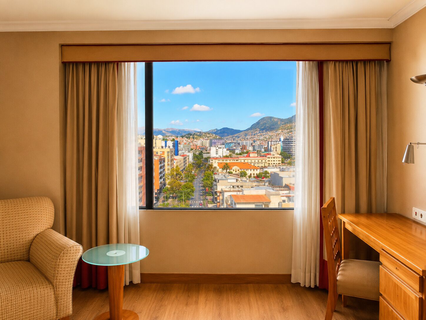 Una habitación de hotel con un ventanal grande, un escritorio de madera, una mesa de vidrio y un sofá a cuadros.