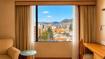 Una habitación de hotel con un ventanal grande, un escritorio de madera, una mesa de vidrio y un sofá a cuadros.