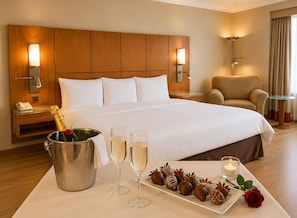 Habitación de hotel con una cama grande, lámparas de noche, un minibar, una mesita de noche con teléfono, una mesa de vidrio con champán, copas y fresas cubiertas de chocolate.