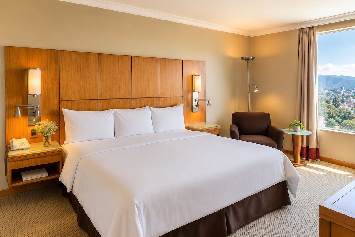 Une chambre d’hôtel avec un grand lit, une tête de lit en bois, des lampes de chevet, une chaise, une petite table et une vue sur la ville par la fenêtre.