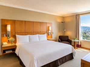 Une chambre d’hôtel avec un grand lit, une tête de lit en bois, des lampes de chevet, une chaise, une petite table et une vue sur la ville par la fenêtre.