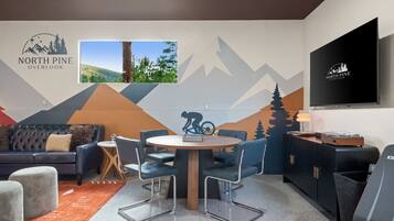 A modern living room with a mountain-themed wall mural, a flat-screen TV, a dining table with chairs, a sofa, and a coffee table.