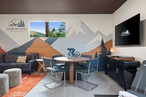 A modern living room with a mountain-themed wall mural, a flat-screen TV, a dining table with chairs, a sofa, and a coffee table.