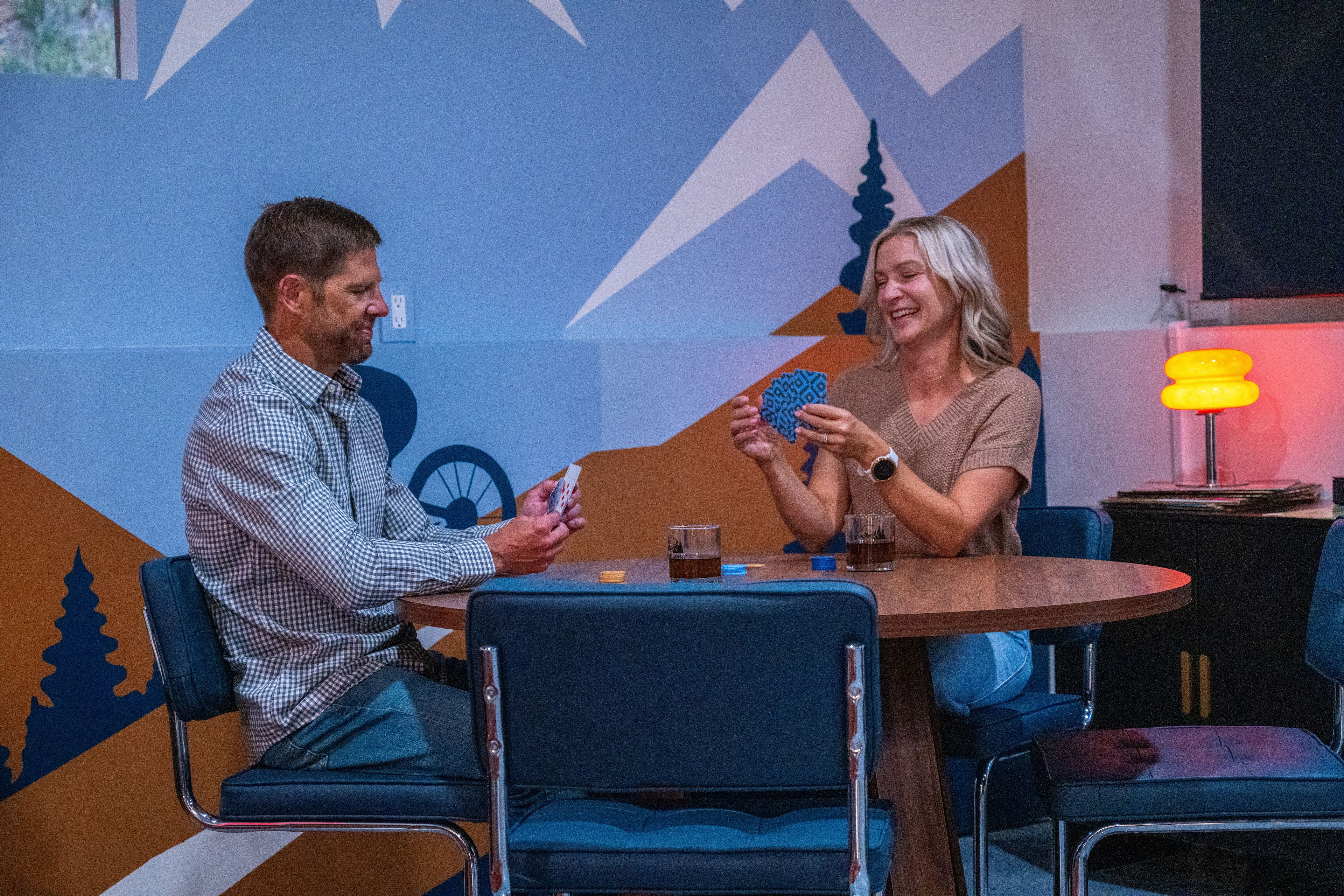 Twee personen zitten aan een ronde tafel, compleet met een blauwe stoel, en spelen kaarten.